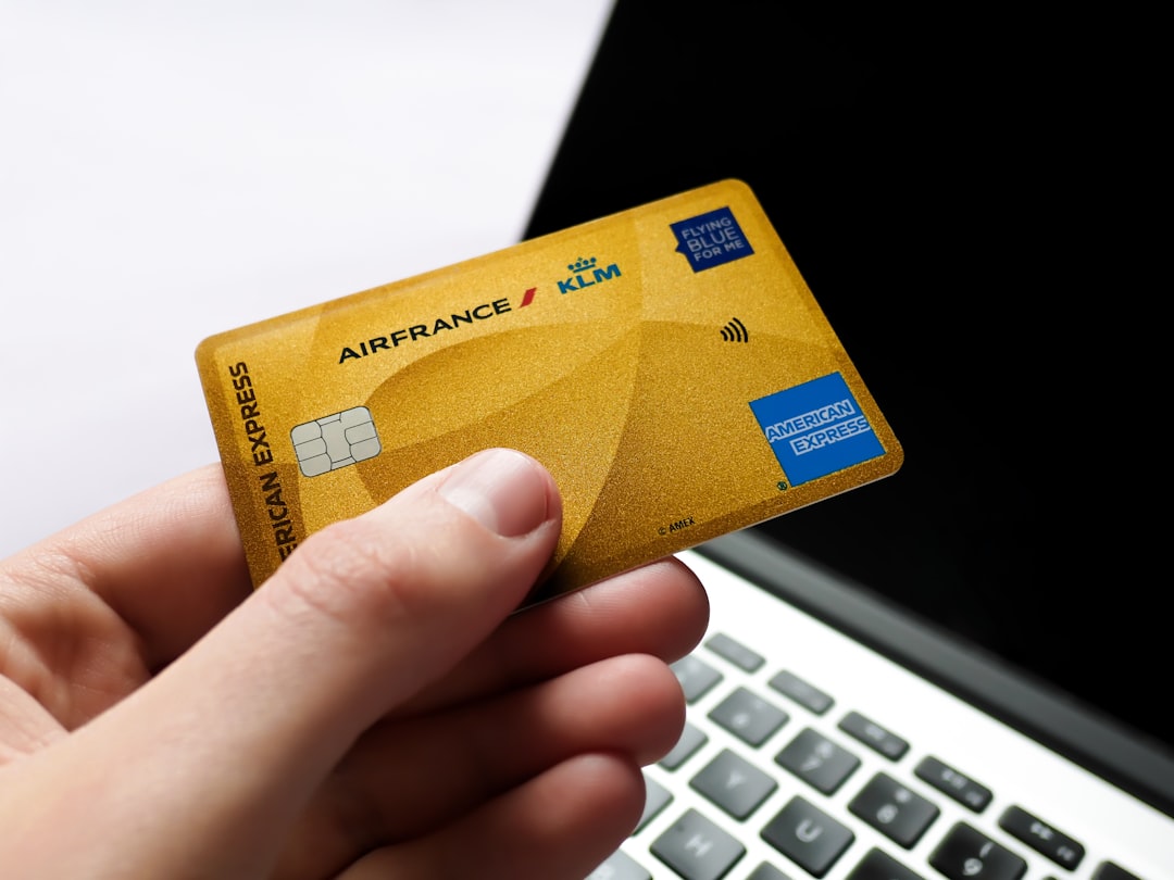 Close-up of a hand holding an American Express credit card with a blurred laptop in the background. This shot captures the essence of finance, online shopping, business travel, and digital payments. The sleek design and subtle Flying Blue branding make it ideal for themes of luxury rewards within the aviation industry or e-commerce loyalty programs.